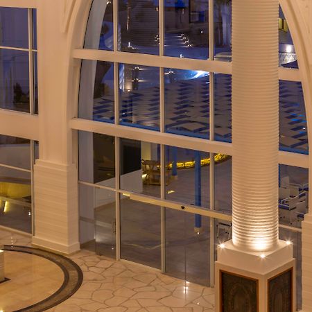 Pickalbatros Palace Sharm - "Aqua Park" Esterno foto The photo shows a spacious interior of a modern building, likely a hotel or a lobby. The scene features large glass windows that allow for a view of areas outside. There are elegant pillars within the space, illuminated from the base, creating a warm