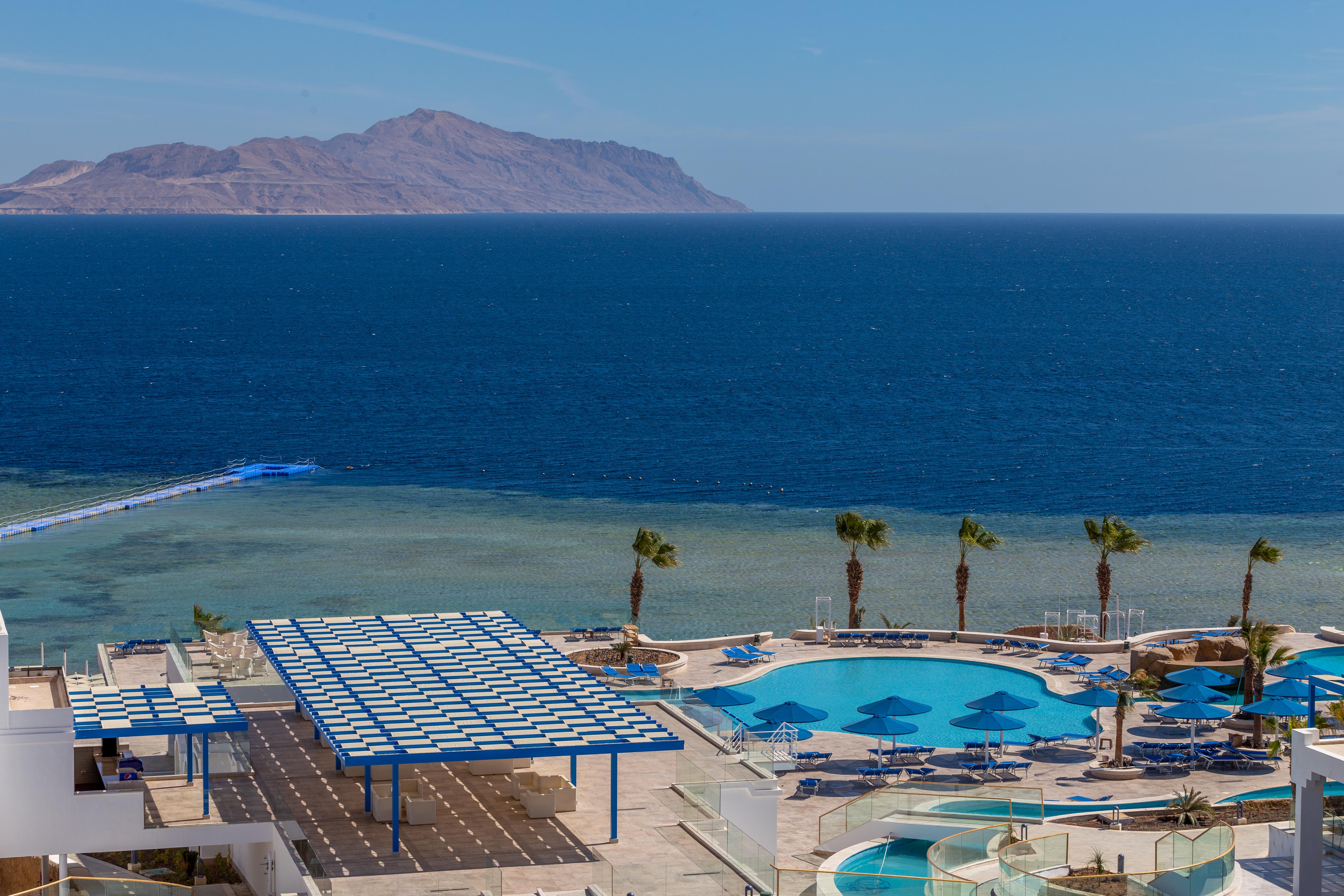 Pickalbatros Palace Sharm - "Aqua Park" Esterno foto The photo shows a beautiful coastal scene featuring a large swimming pool area with lounge chairs and umbrellas. In the background, there is a clear blue ocean stretching out to the horizon, with a distant landmass or mountain visible. Palm trees are