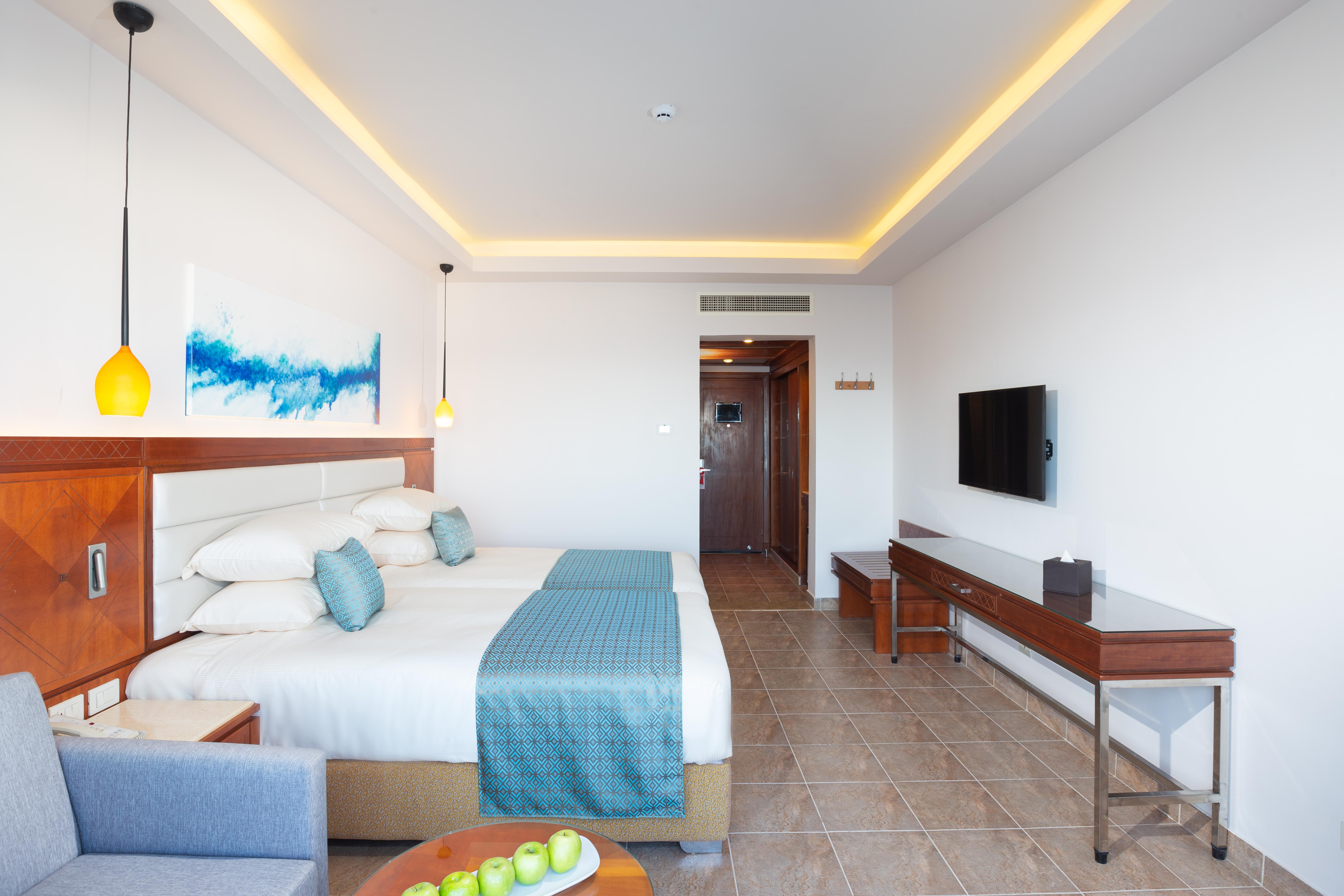 Pickalbatros Palace Sharm - "Aqua Park" Esterno foto The photo shows a modern hotel room featuring two queen-sized beds with white linens and a light blue throw. The room is well-lit, with a warm yellow glow from the ceiling lights. There is a painting on the wall above the beds, characterized by blue 