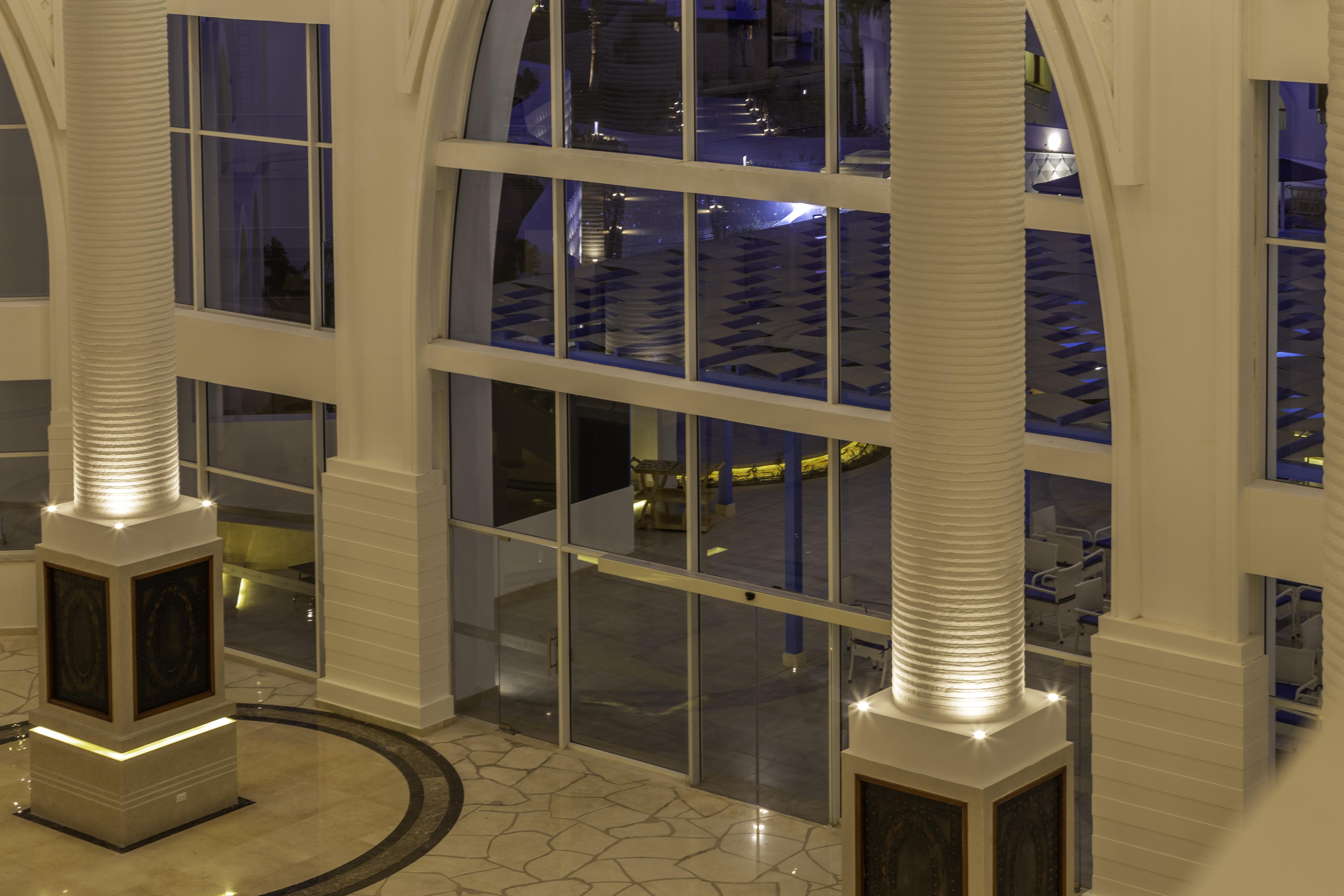 Pickalbatros Palace Sharm - "Aqua Park" Esterno foto The photo shows a spacious interior of a modern building, likely a hotel or a lobby. The scene features large glass windows that allow for a view of areas outside. There are elegant pillars within the space, illuminated from the base, creating a warm