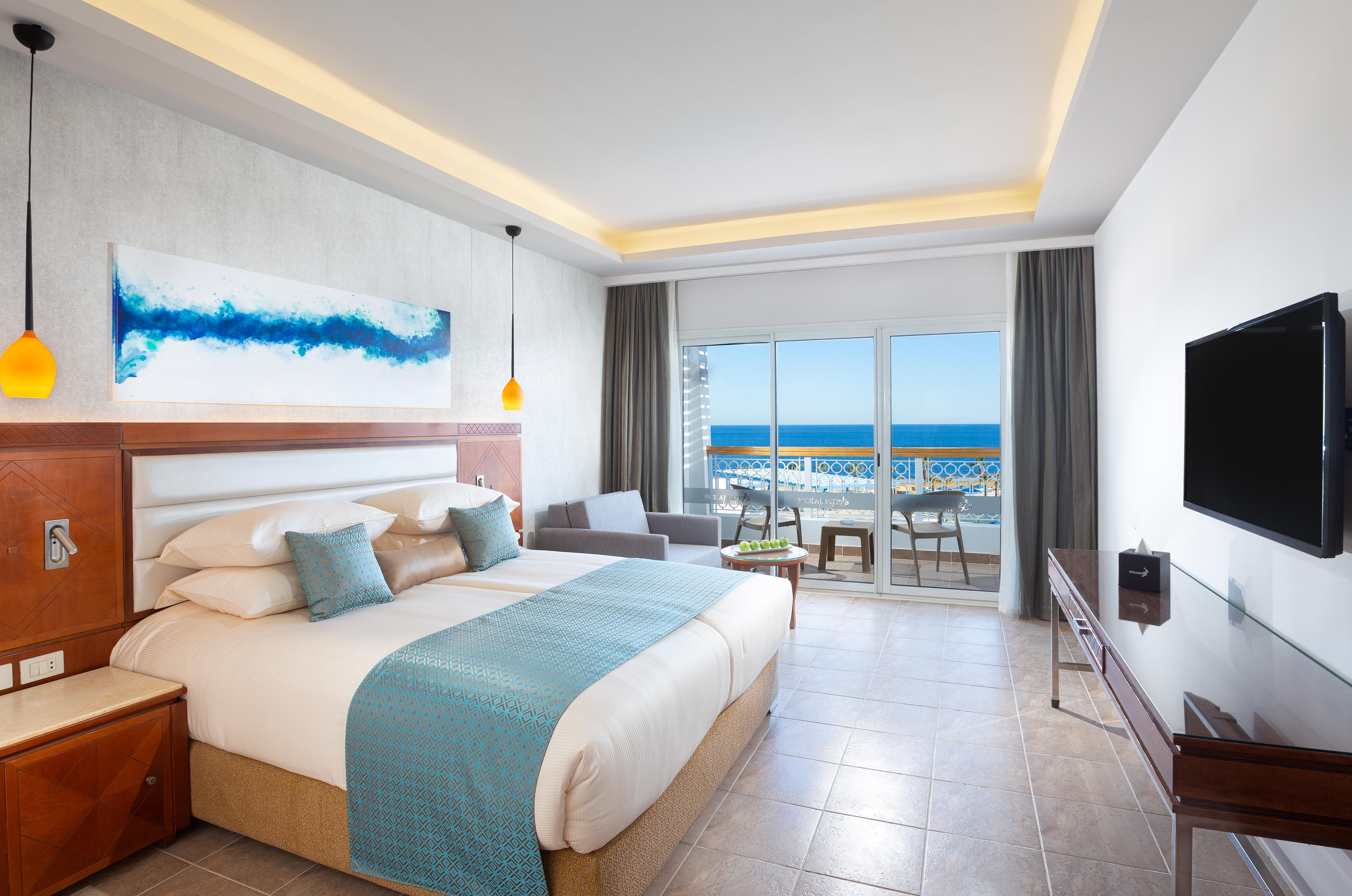 Pickalbatros Palace Sharm - "Aqua Park" Esterno foto The photo shows a modern hotel room with a large bed dressed in white linens and a light blue throw blanket. There is a piece of abstract artwork on the wall above the bed, featuring blue tones. The room has large windows with a view of the ocean, al