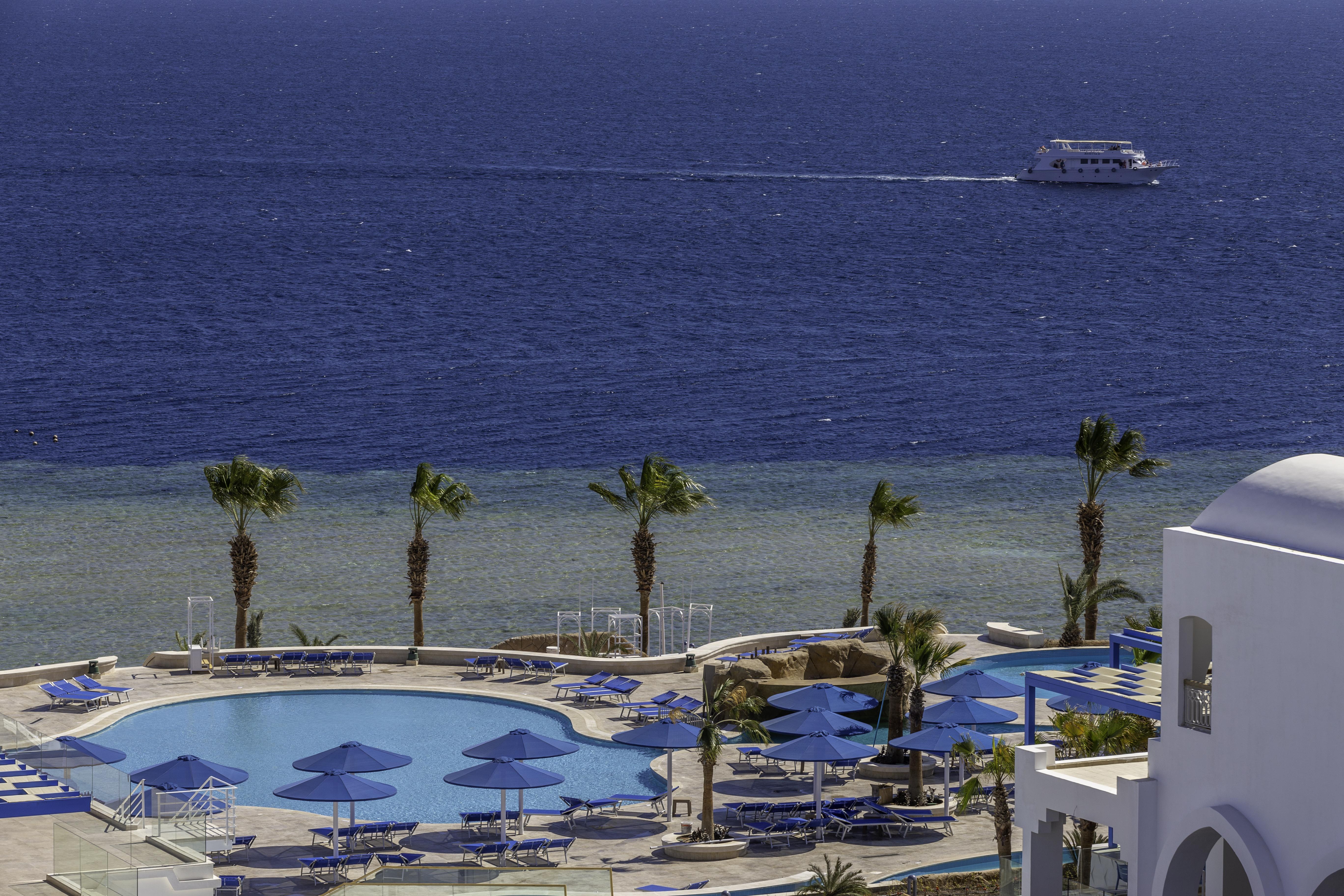 Pickalbatros Palace Sharm - "Aqua Park" Esterno foto The photo depicts a serene seaside view featuring a large swimming pool surrounded by lounge chairs and umbrellas. In the background, there is a calm sea with a boat cruising on the water. Palm trees line the edge of the pool area, adding to the trop