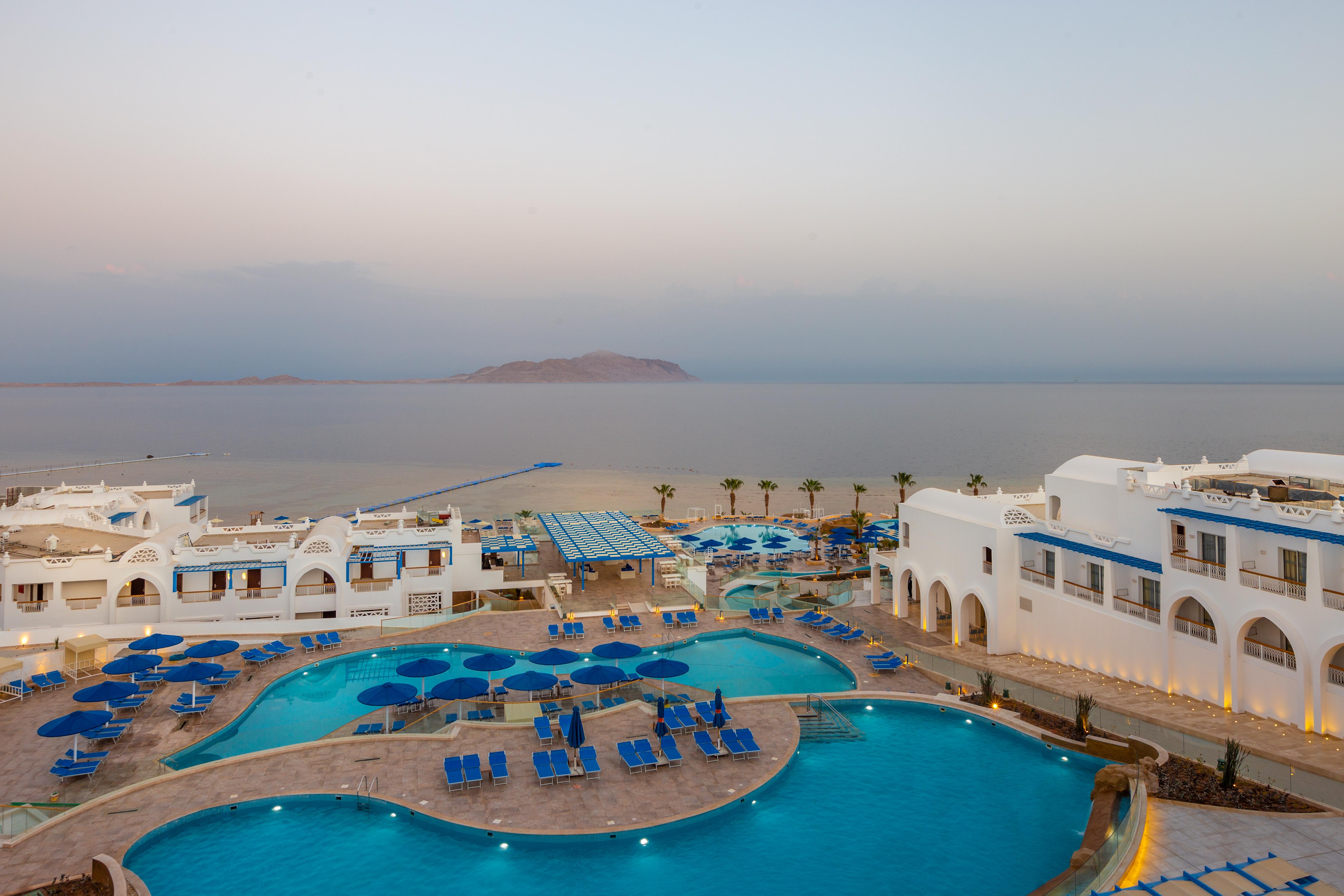 Pickalbatros Palace Sharm - "Aqua Park" Esterno foto The photo shows a resort area with expansive pools surrounded by lounge chairs. The pools are designed in a curvy shape, and there are numerous blue umbrellas scattered around. In the background, there is a calm sea stretching out towards a distant l