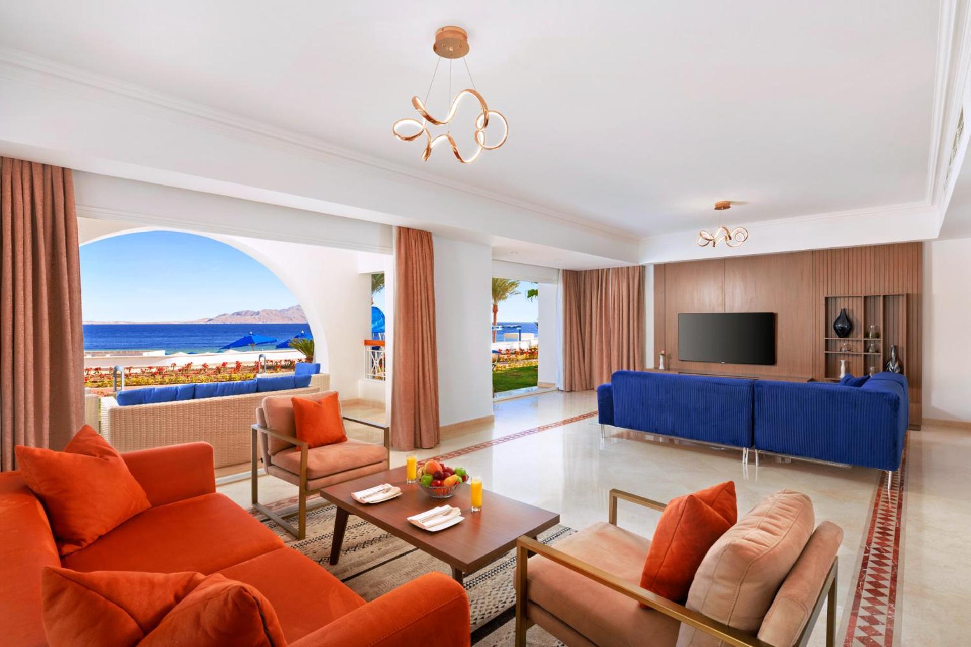 Pickalbatros Palace Sharm - "Aqua Park" Esterno foto The photo showcases a stylish and modern living space that features a bright and open layout. The room includes a cozy seating area with orange couches and a wooden coffee table, adorned with a fruit arrangement. Large windows provide a view of the o