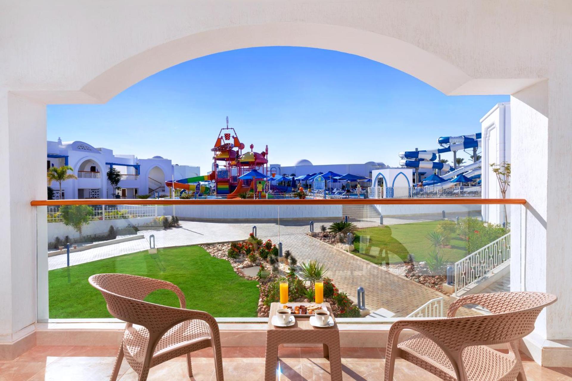 Pickalbatros Palace Sharm - "Aqua Park" Esterno foto The photo shows a view from a balcony with two chairs and a small table set up for relaxation. In the foreground, there is a breakfast setup with orange juice and a pastry. Beyond the balcony, there is a vibrant, colorful water play area with slides 