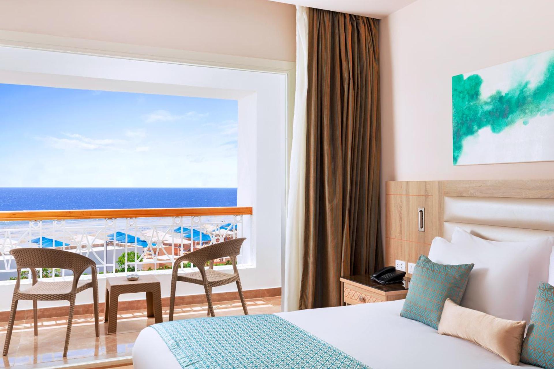 Pickalbatros Palace Sharm - "Aqua Park" Esterno foto The photo shows a modern hotel room featuring a large bed with white linens and a teal accent pillow. There is a window with a view of the ocean, framed by light curtains, which lets in natural light. In front of the window, there are two wooden chai