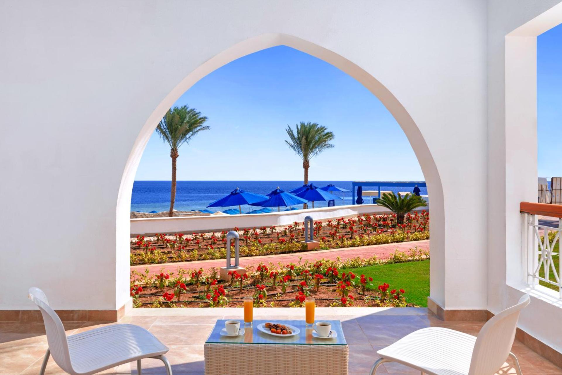 Pickalbatros Palace Sharm - "Aqua Park" Esterno foto The photo depicts a serene view through an architectural archway. In the foreground, there is a small table set with two glasses of orange juice and a plate of snacks. Surrounding the table are two white chairs. Beyond the arch, you can see a calm bl