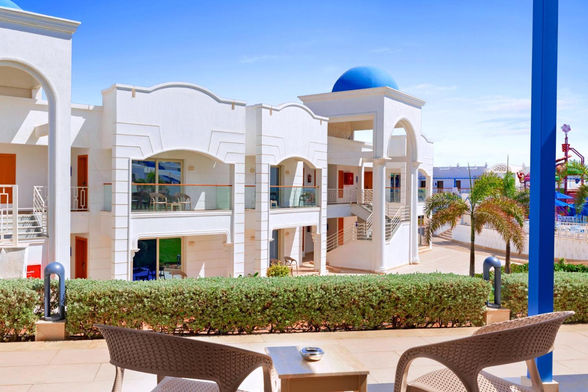 Pickalbatros Palace Sharm - "Aqua Park" Esterno foto The photo shows a modern architectural setting, likely at a resort or hotel. It features white buildings with decorative elements and balconies. The scene is bright with blue skies, and there are palm trees visible, adding a tropical vibe. In the for