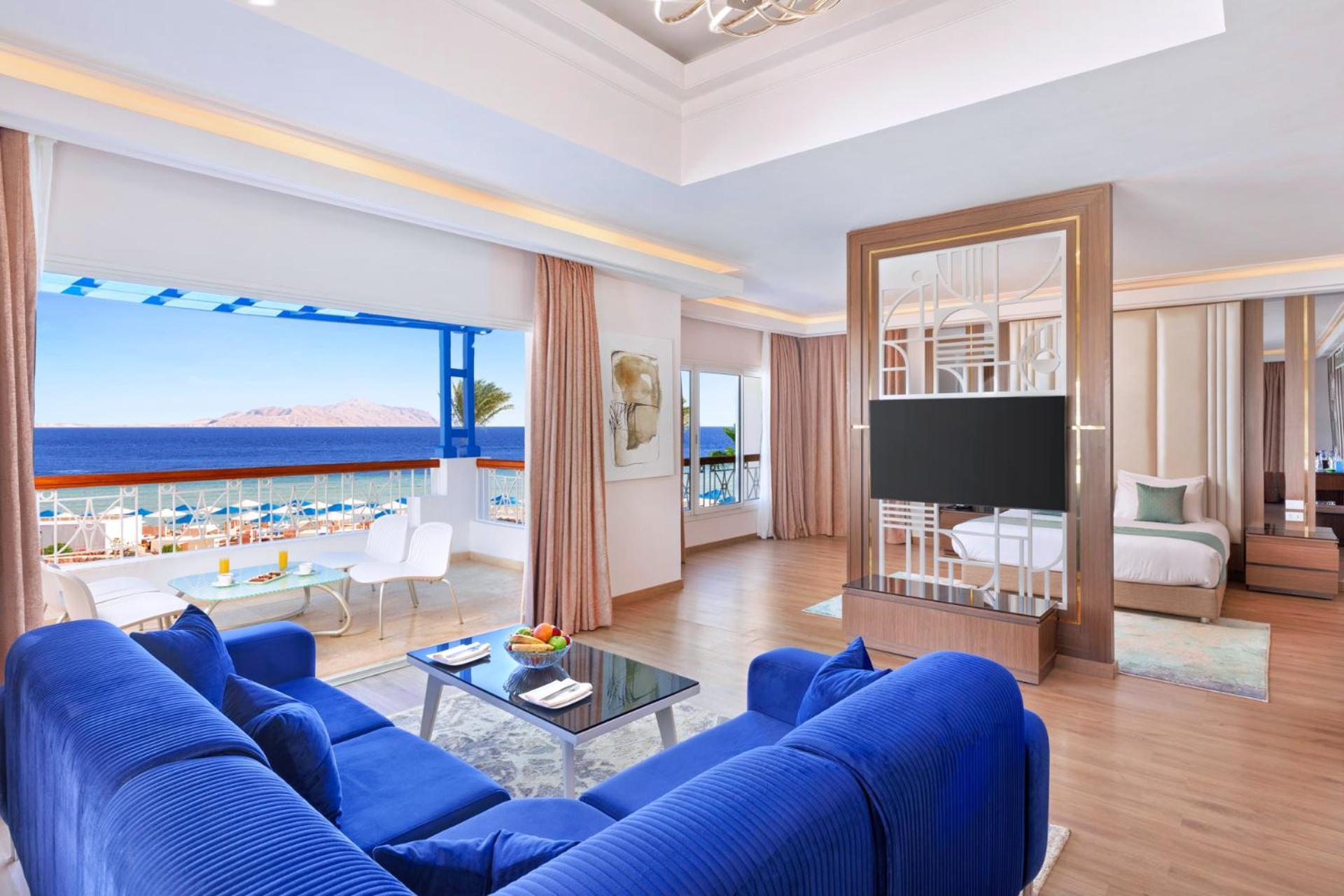 Pickalbatros Palace Sharm - "Aqua Park" Esterno foto The photo shows a well-appointed hotel room or suite featuring a modern and stylish design. There is a spacious living area with a large blue sofa and a glass coffee table in the center. The room is bright, with natural light coming in from large win