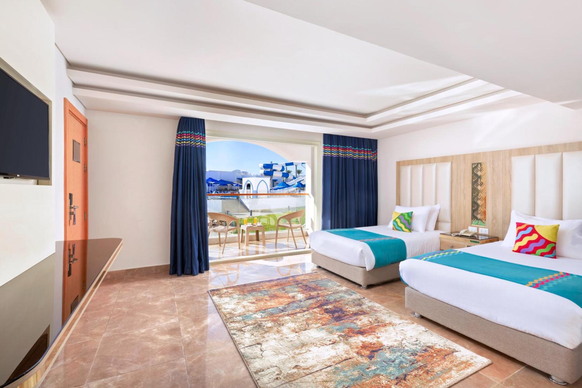 Pickalbatros Palace Sharm - "Aqua Park" Esterno foto The photo shows a well-decorated hotel room featuring two beds, each with colorful accent pillows. The walls are painted in a light color, and there is a large window with blue curtains that offers a view of an outdoor area. A small seating area with