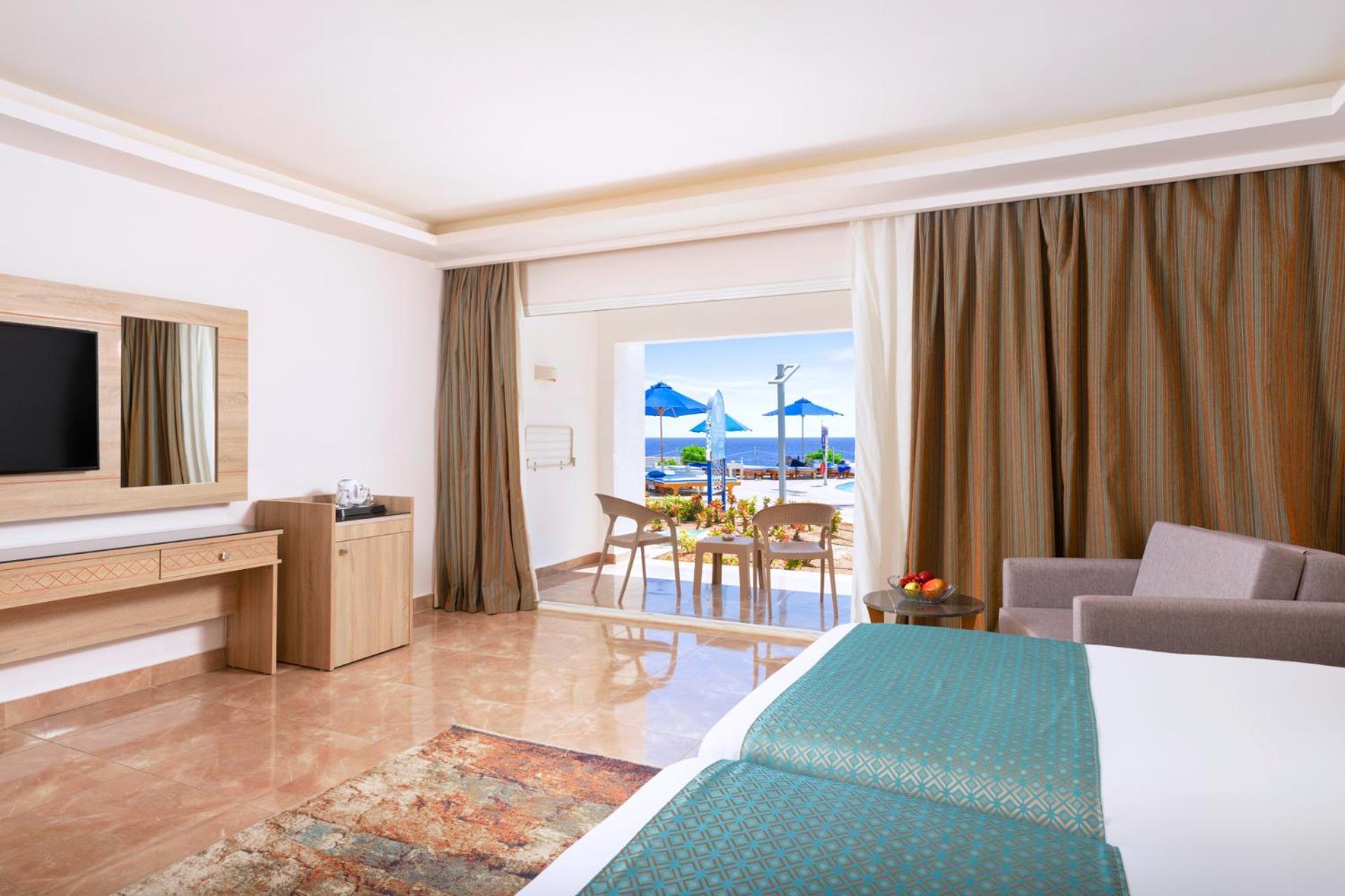 Pickalbatros Palace Sharm - "Aqua Park" Esterno foto The photo shows a spacious hotel room designed with a modern and elegant style. It features two beds with teal-colored bedding, a large mirror, and a television on the wall. There are wooden furniture pieces, including a dresser and a small table. To