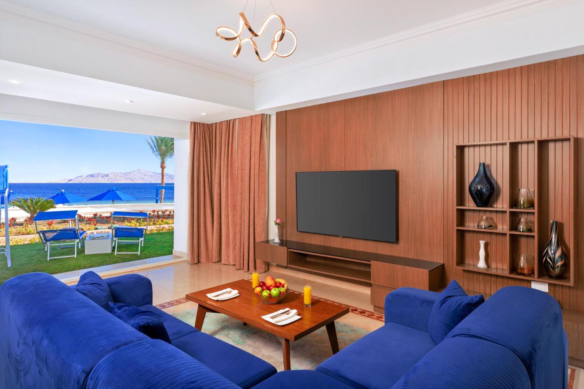 Pickalbatros Palace Sharm - "Aqua Park" Esterno foto The photo shows a modern living room with a stylish design. It features a blue sofa arranged around a wooden coffee table, which has some decorative items, including fruit and candles. A large flat-screen TV is mounted on a wooden wall panel. There a