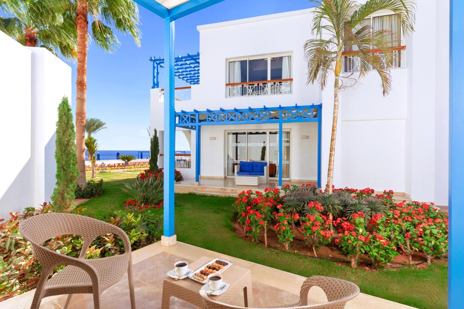 Pickalbatros Palace Sharm - "Aqua Park" Esterno foto The photo shows a picturesque outdoor setting, likely part of a resort or hotel. In the foreground, there are two stylish chairs and a small table with two cups, possibly of coffee or tea, along with a plate of snacks. The background features a well-