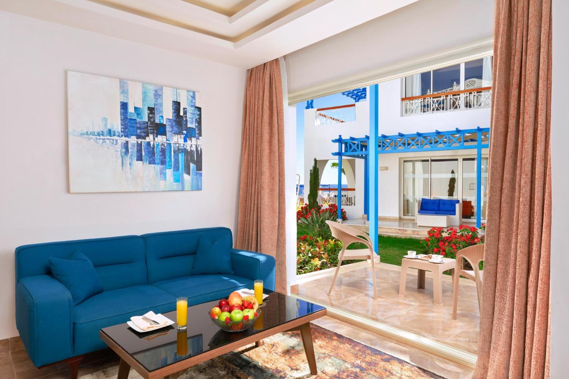Pickalbatros Palace Sharm - "Aqua Park" Esterno foto The photo shows a bright and modern living space. There is a blue couch facing a glass door that leads to a garden area. In front of the couch, there's a dark coffee table with some fruit and candles on it. A large piece of abstract artwork in blues 