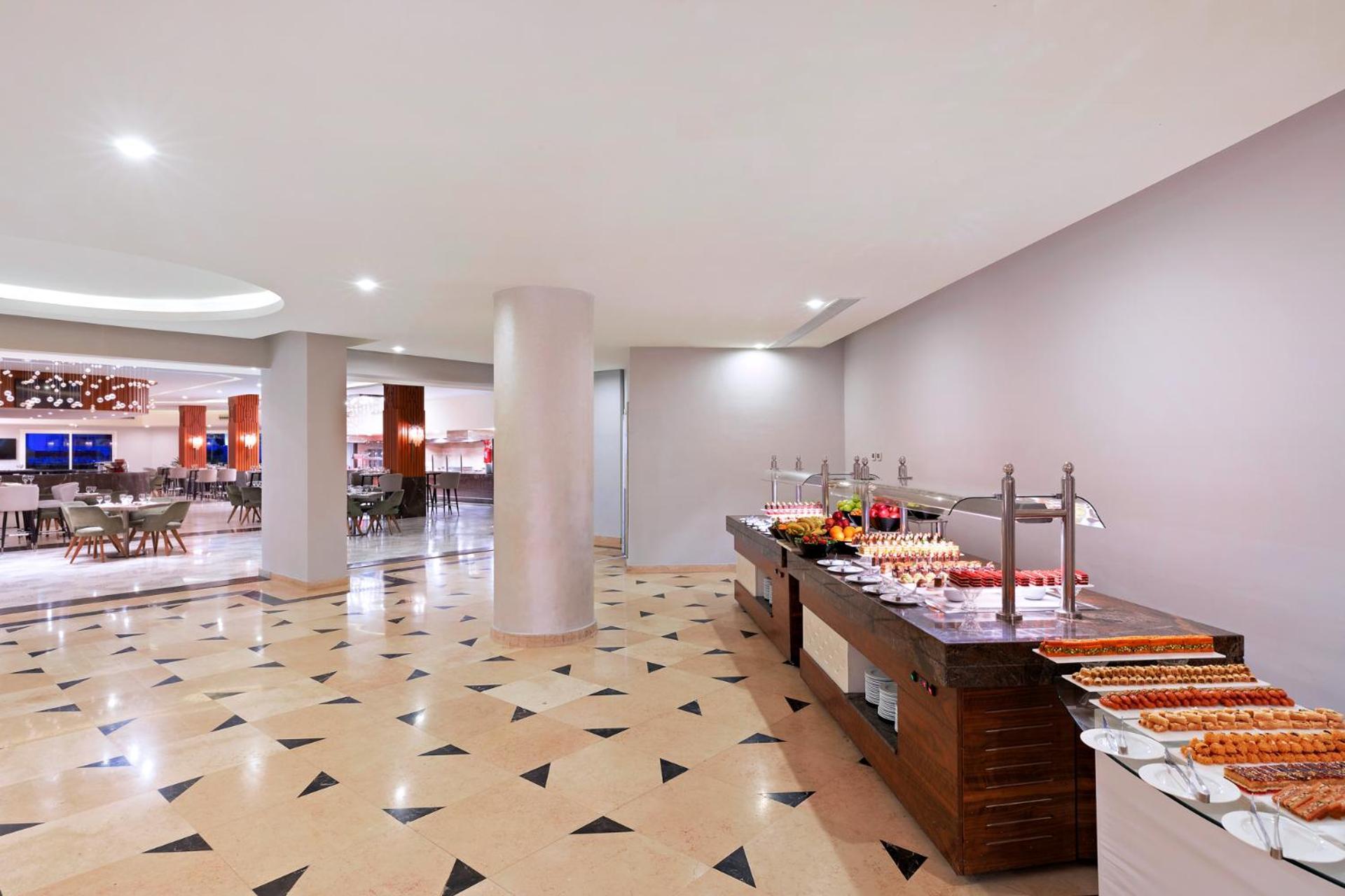 Pickalbatros Palace Sharm - "Aqua Park" Esterno foto The photo shows a modern dining area, likely a buffet or restaurant space. In the foreground, there's a wooden buffet table displaying various food items, including desserts. The floor has a decorative tile pattern, featuring a mix of light and darke