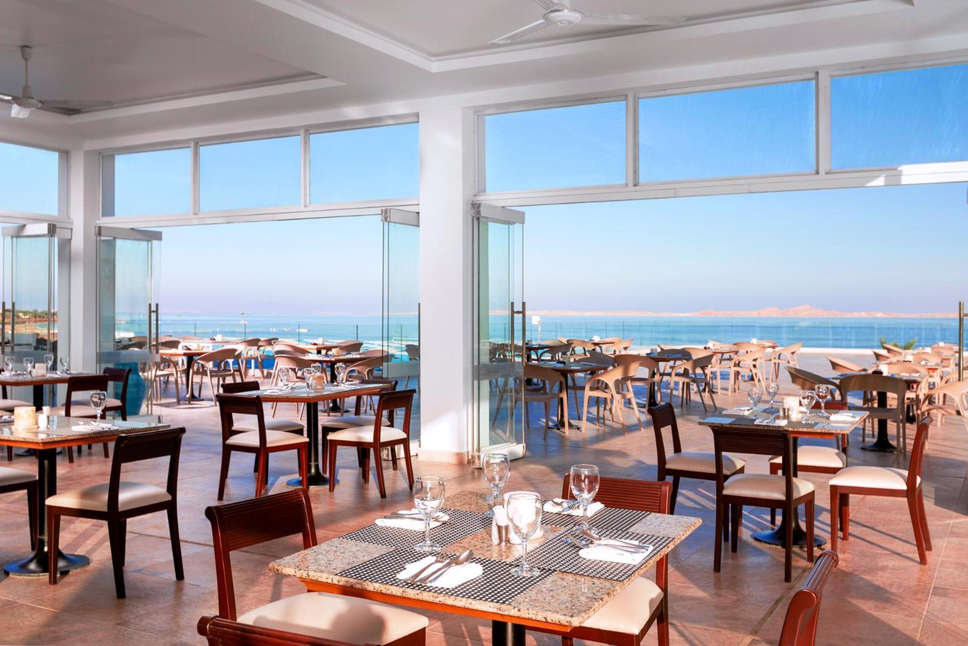 Pickalbatros Palace Sharm - "Aqua Park" Esterno foto The photo shows a bright and modern restaurant with large windows that offer a scenic view of the sea. The interior features wooden tables and chairs, neatly arranged for dining. The atmosphere appears open and airy, thanks to the ample natural light